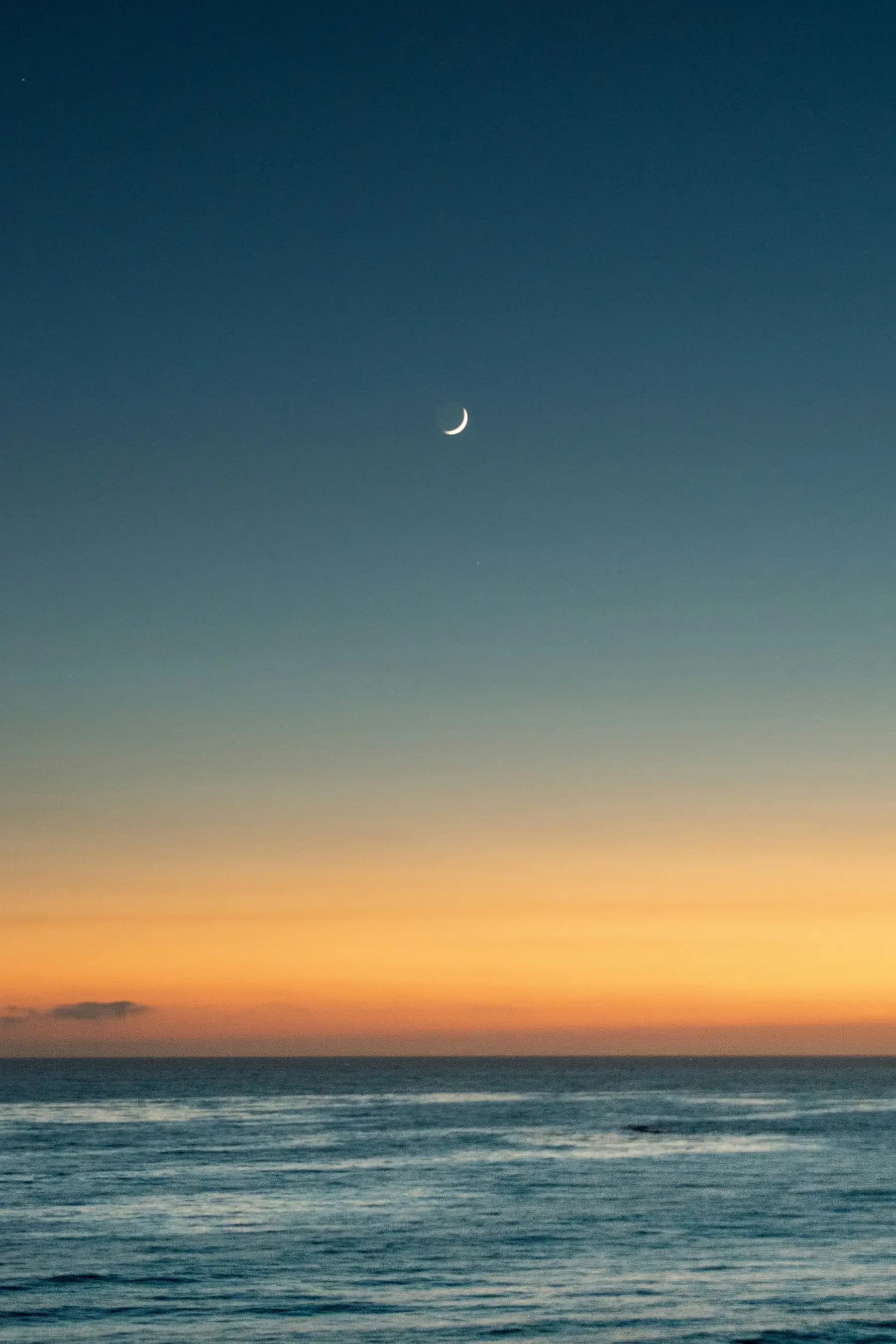 A crescent moon in a clear sky above the ocean at dusk