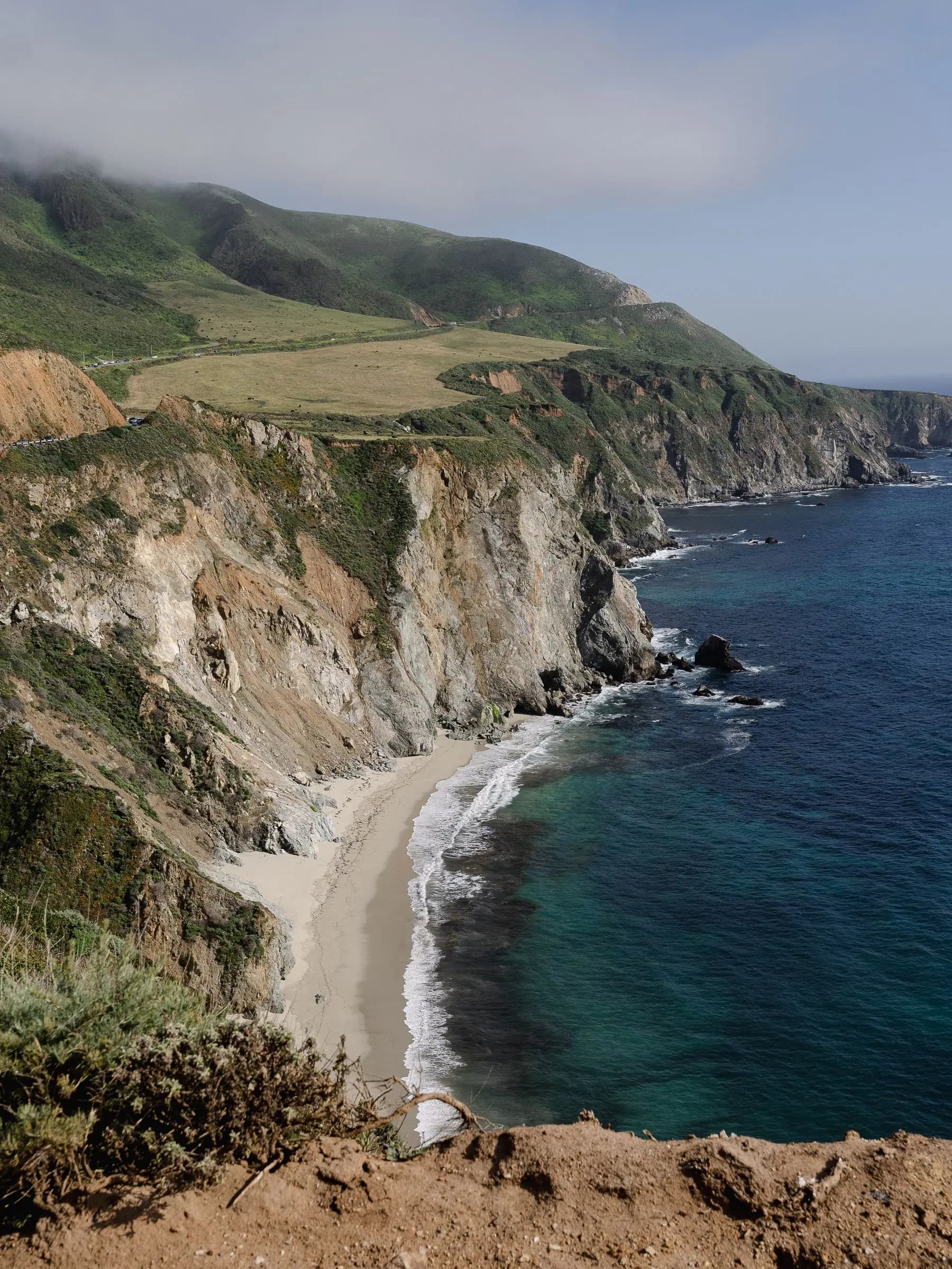 A coastal road and bridge along green cliffs above a turquoise cove